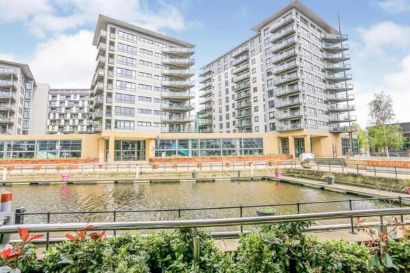 A modern residential building with multiple balconies, a central courtyard with water features, and a clear sky.