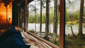 Una cabaña con cama, ventana y vistas a un bosque y un río.