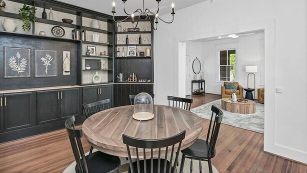Un'area pranzo con un tavolo rotondo, sedie e un lampadario. Si nota una libreria con diversi oggetti e una porta che dà su un'altra stanza.