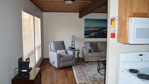 Un espacio habitable compacto con techo de madera, estufa blanca, sillón gris, sofá, una mesita con lámpara y un cuadro en la pared.