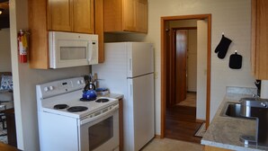 Cocina con armarios de madera, estufa blanca, microondas y refrigerador. Hay un lavamanos y una puerta que da a otra habitación.