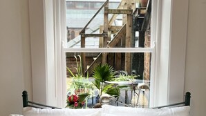 Un lit avec une literie blanche et deux oreillers verts, une fenêtre donnant sur un escalier en bois et des plantes à l’extérieur.