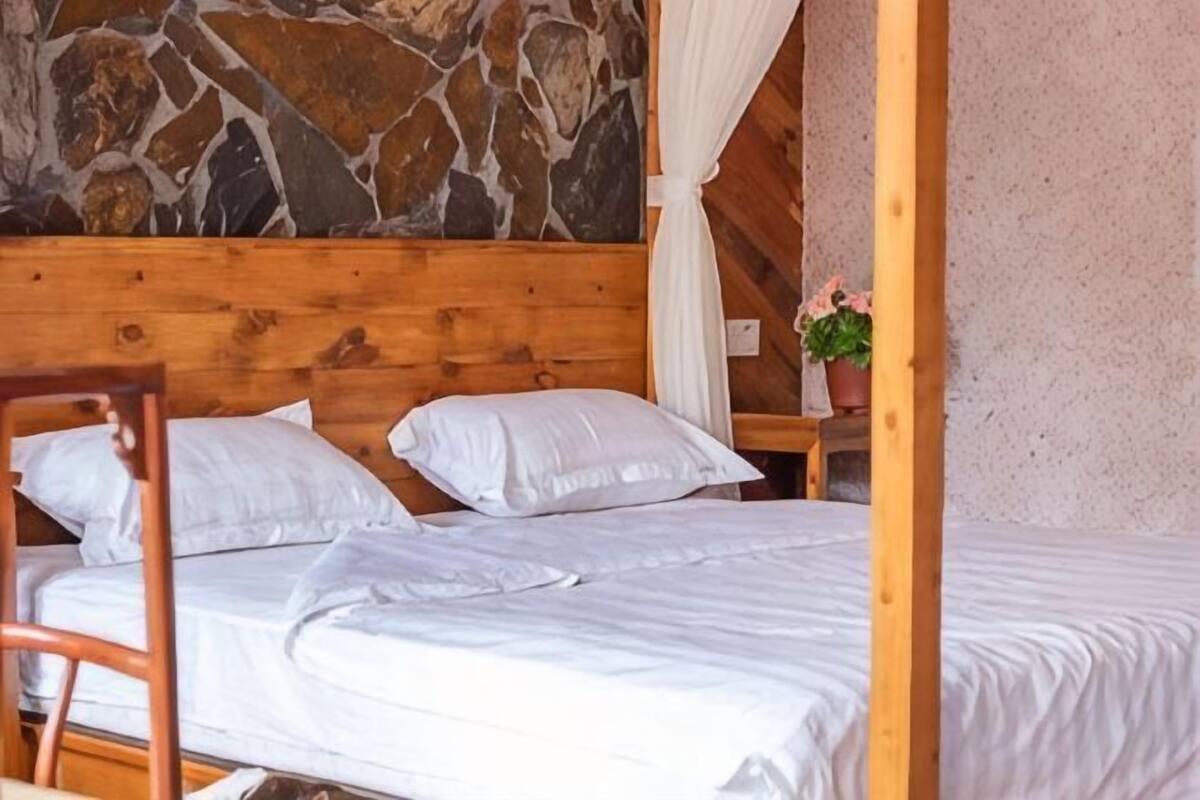 A wooden bed with white bedding, a stone wall, and a small table with a potted plant.
