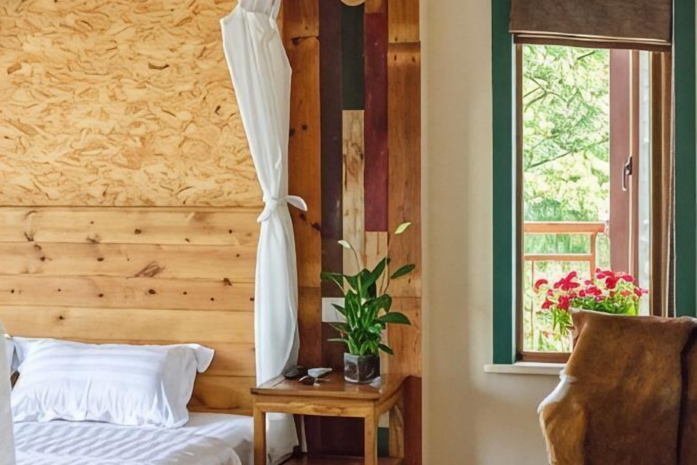 A wooden room with a bed, a small table with a plant, a chair, and a window with a view of greenery.