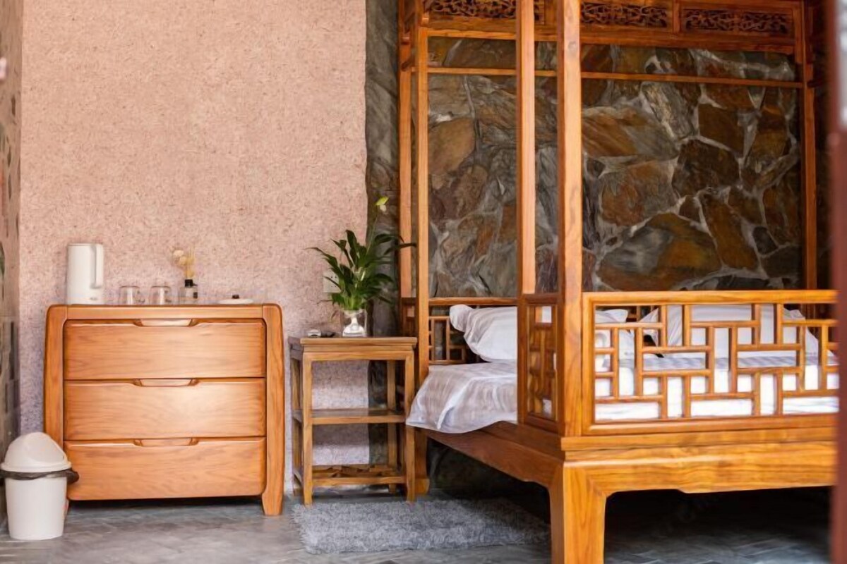 A bedroom with a wooden bed, a nightstand, a dresser, and a stone wall.
