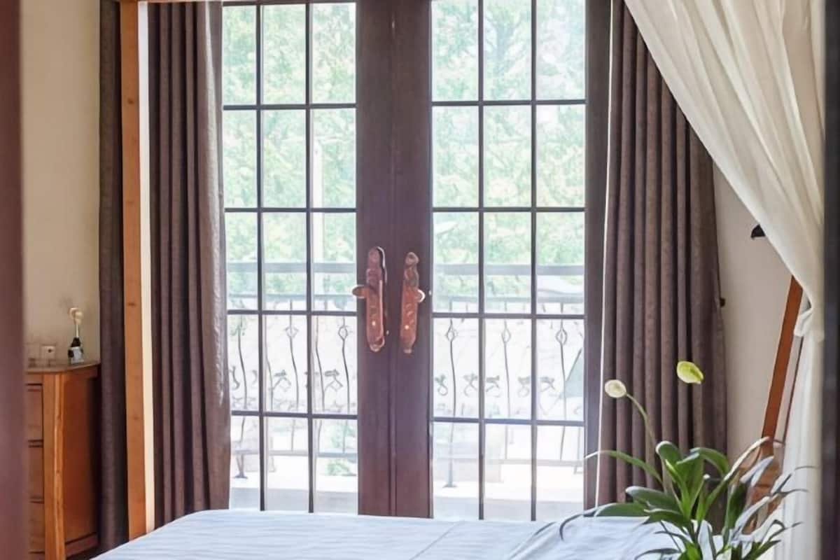 A room with a large window, curtains, a bed, a wooden dresser, and a potted plant.