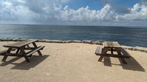 Una playa con dos mesas de picnic de madera y un muro de piedra al fondo.
