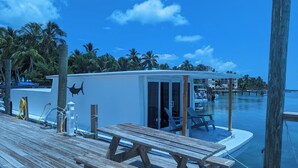 Un muelle de madera con un área de estar cubierta y un pequeño edificio con puerta, con vista a una masa de agua tranquila con barcos y palmeras al fondo.