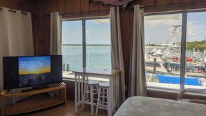 Una habitación con techo de madera, un televisor sobre un soporte de madera, una mesa con dos sillas blancas, un ventanal con cortinas y un pez montado en la pared.