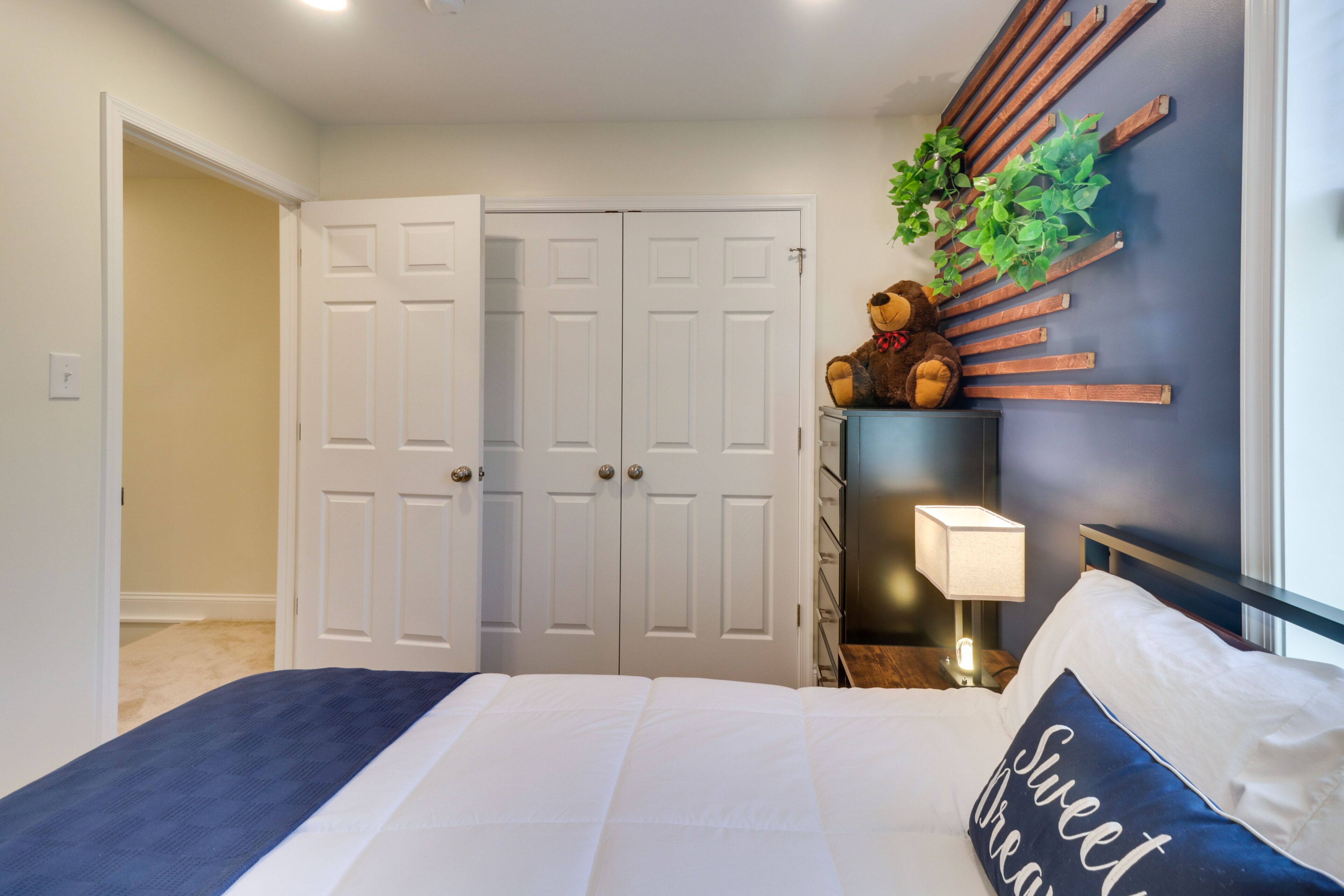 A bedroom with a bed, a nightstand, a lamp, a teddy bear, and a wall-mounted plant.