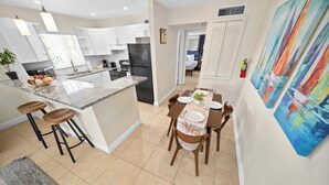 Una cocina moderna con armarios blancos, una isla de mármol y una zona de comedor con sillas y mesa de madera.