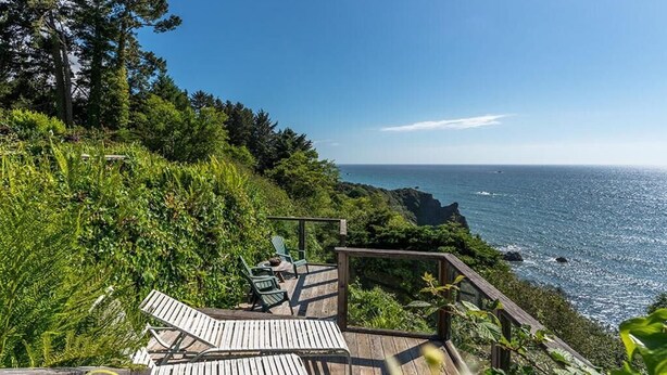 Une terrasse en bois, agrémentée de chaises, qui offre une vue sur une zone côtière caractérisée par des affleurements rocheux et une végétation luxuriante.