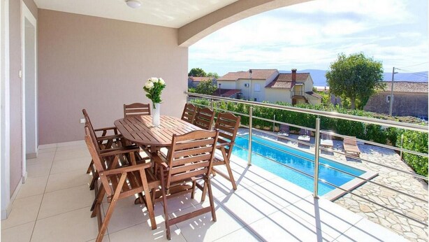 Un balcón con una mesa y sillas de madera, un pequeño jarrón blanco con flores, y una vista de una piscina y casas al fondo.