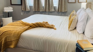 A neatly made bed with a yellow throw, a bedside table with books, and a window with curtains. - Walk to ACRISURE in 23 Mins!  King Bed + FREE private park ✭Perfect Locale✭ (Pittsburgh)
