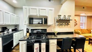 A kitchen with white cabinets, a stove, a microwave, and a coffee maker. There is a bar area with two chairs and a small table. - Walk to ACRISURE in 23 Mins!  King Bed + FREE private park ✭Perfect Locale✭ (Pittsburgh)