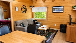 A cozy cabin interior with a wooden dining table, a sofa, a small table with a book, a TV on a wooden cabinet, and a window with a view of greenery. - Box Canyon Cabin Bear's Den Cabin - 2 bed, WI-FI (Seward)