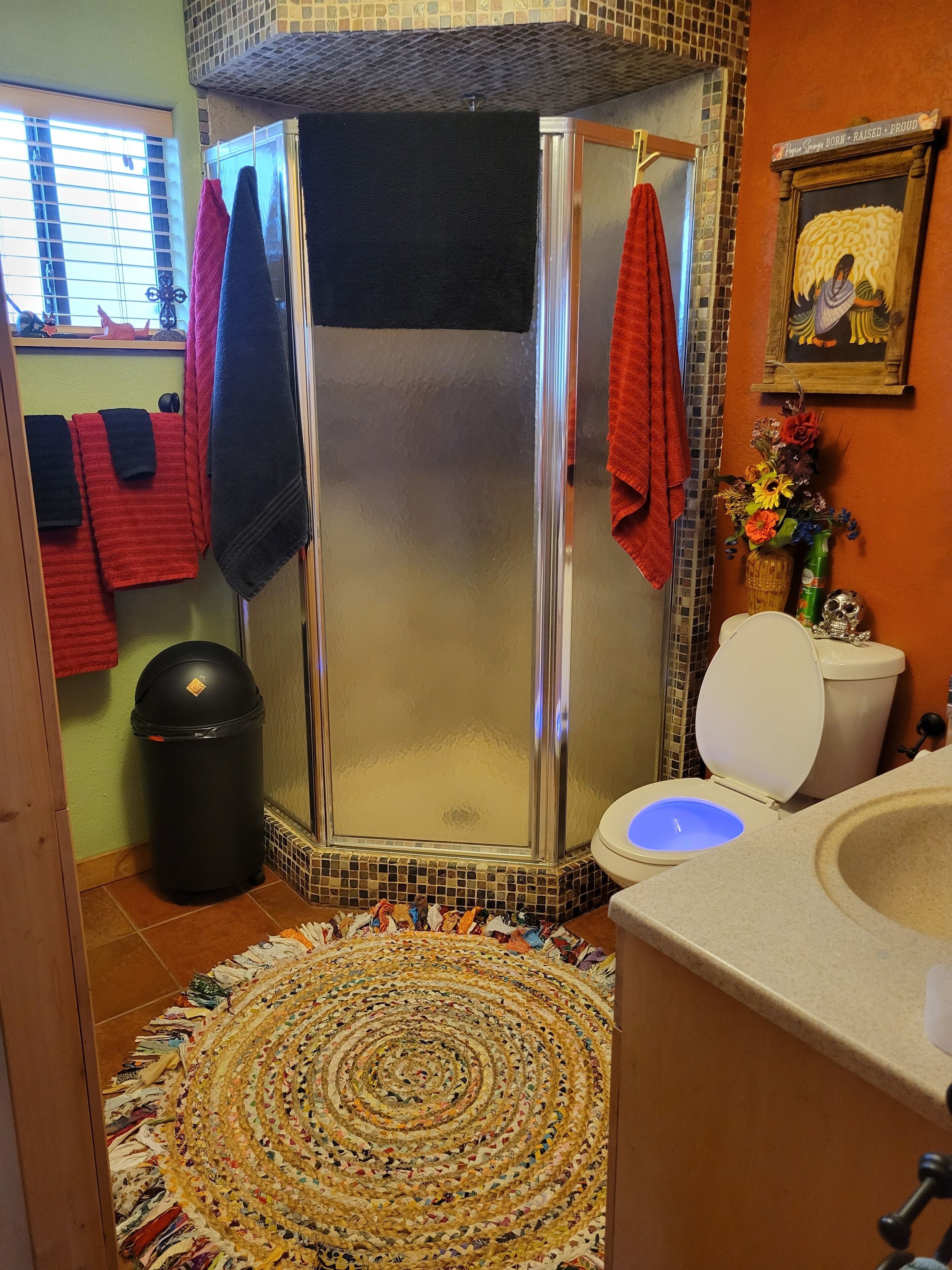 A bathroom with a glass-enclosed shower, a toilet with a blue-lit bowl, a trash bin, and a patterned rug on the floor.