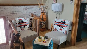 Ein gemütliches Wohnzimmer mit zwei grauen Sesseln, einem Holztisch, einem roten Teppich, einer Holzkommode, einer Vase mit Zweigen und einer Lampe.