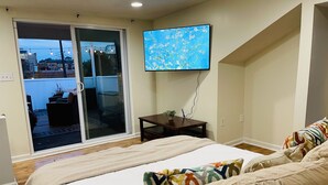 Un dormitorio con una cama, un televisor montado en la pared, una mesita con una planta y un ventanal corredizo que da a un espacio exterior.