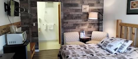 A bedroom with a bed, two chairs, a bedside table, a lamp, a microwave, and a wooden wall panel.