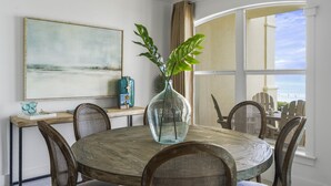 A dining area with a round wooden table, wicker chairs, a glass vase with a plant, and a large abstract painting on the wall. - SPECTACULAR CONDO, GULF-FRONT, VIEWS, POOL, GRILL, LG. PATIO, PRIVATE BEACH (Santa Rosa Beach)
