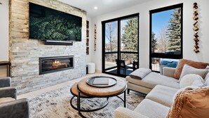 Un salon moderne avec une cheminée en pierre, un téléviseur à écran plat fixé au mur, un ventilateur de plafond et de grandes fenêtres donnant sur la forêt.