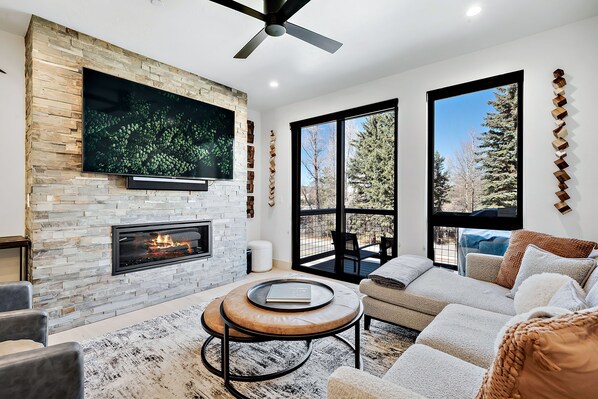 A modern living room with a stone fireplace, a flat-screen TV mounted on the wall, a ceiling fan, and large windows offering a view of trees.