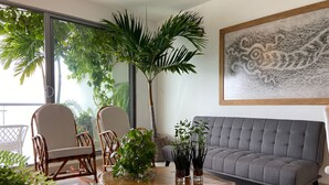 Un salon moderne avec un canapé gris, deux fauteuils en osier, une table basse en bois agrémentée de plantes et une grande fenêtre donnant sur un paysage verdoyant.