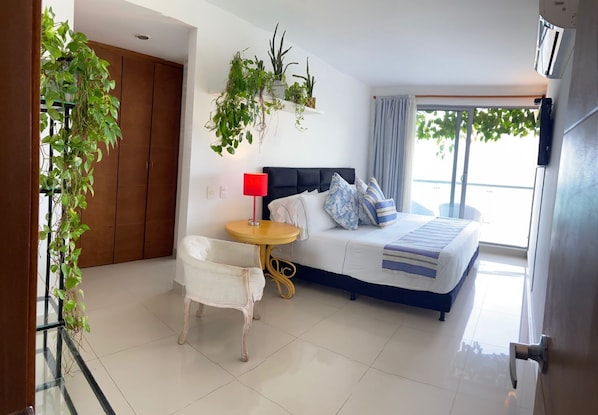 A modern hotel room with a bed, a small table with a lamp, a chair, and potted plants.