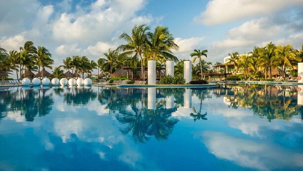 Une piscine aux eaux cristallines, bordée de palmiers et de chaises longues, le tout intégré dans un cadre de station balnéaire tropical.