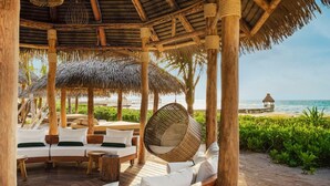 Une terrasse en bois surmontée d’un toit de chaume, dotée de sièges blancs rembourrés et d’un fauteuil suspendu en osier.