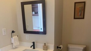 Un baño con lavamanos blanco, tocador de madera oscura, un espejo, inodoro y una cesta.