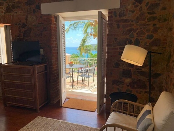 A room with a stone wall, a wooden dresser, a TV, a lamp, and a view of the ocean through an open door. - La Terazza w/ Queen Bed (Charlotte Amalie)