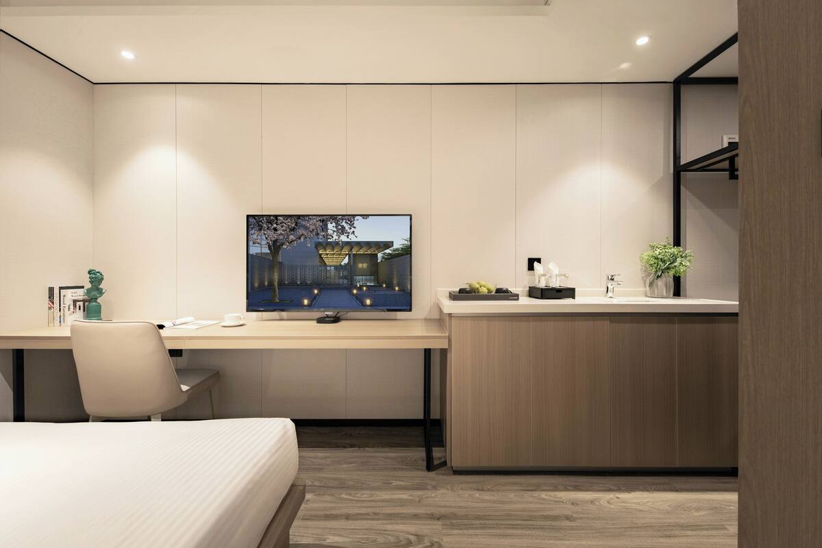 A modern hotel room with a bed, a desk with a computer, a TV, and a small kitchenette.