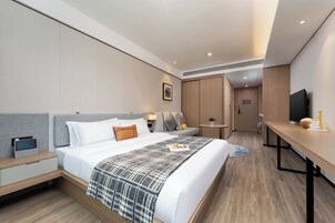 A modern hotel room with a neatly made bed, bedside table with a radio, a small table with a lamp, a TV on a stand, and a door leading to a bathroom.