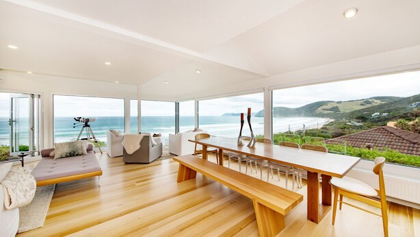 Una habitación amplia con piso de madera, una gran mesa de comedor y un sofá. Se observa un telescopio cerca de la ventana, que ofrece vistas al océano.