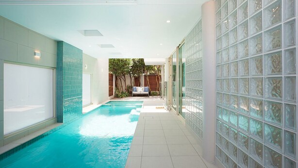 A modern indoor swimming pool with clear glass walls, a tiled floor, and a seating area with a sofa and cushions. - Gordons Bay Vista (Clovelly)