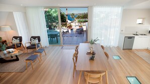 A modern living room with a wooden dining table, comfortable seating, a kitchen area, and a balcony with a view of the sea. - Gordons Bay Vista (Clovelly)