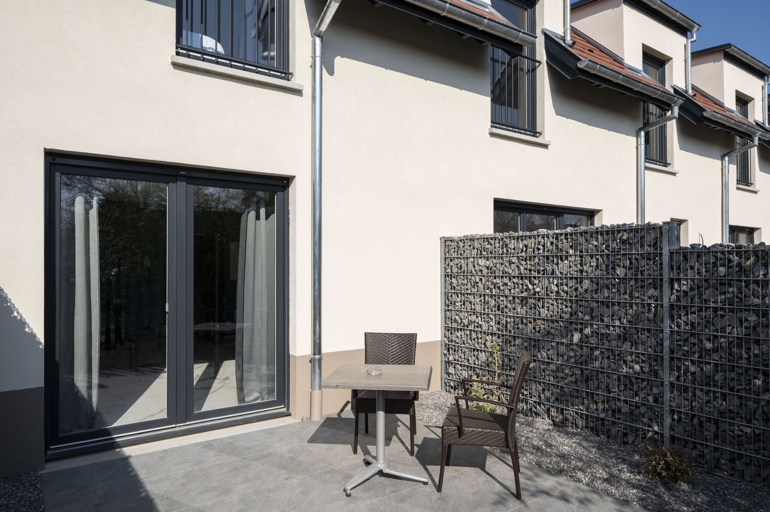 Un edificio moderno con un gran ventanal corredera, una pequeña mesa con sillas y una pared de piedra.