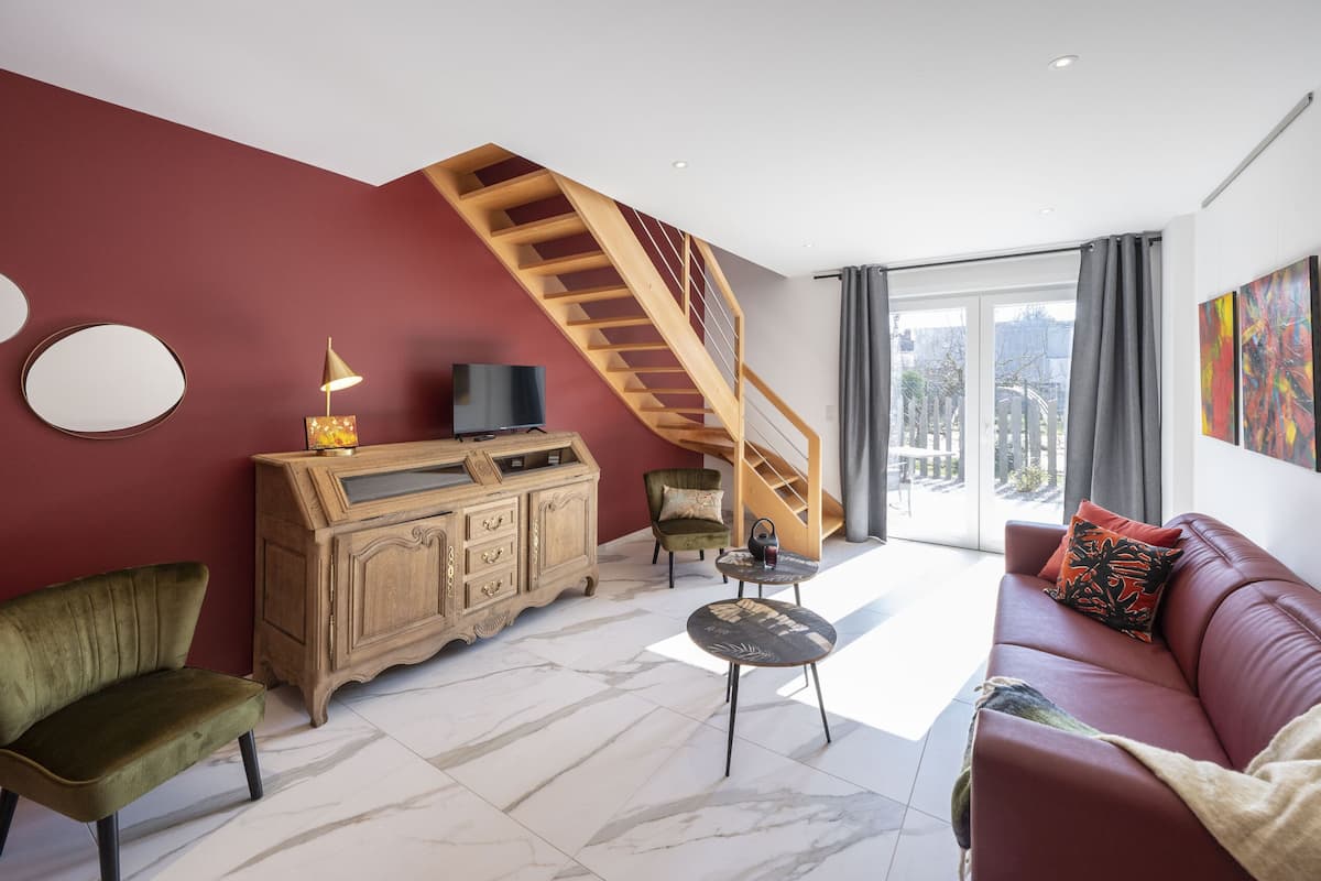 Ein Wohnzimmer mit roter Wand, einer Holztreppe, einem Ledersofa, einem grünen Sessel, einem Holzkasten und einem Fernseher.