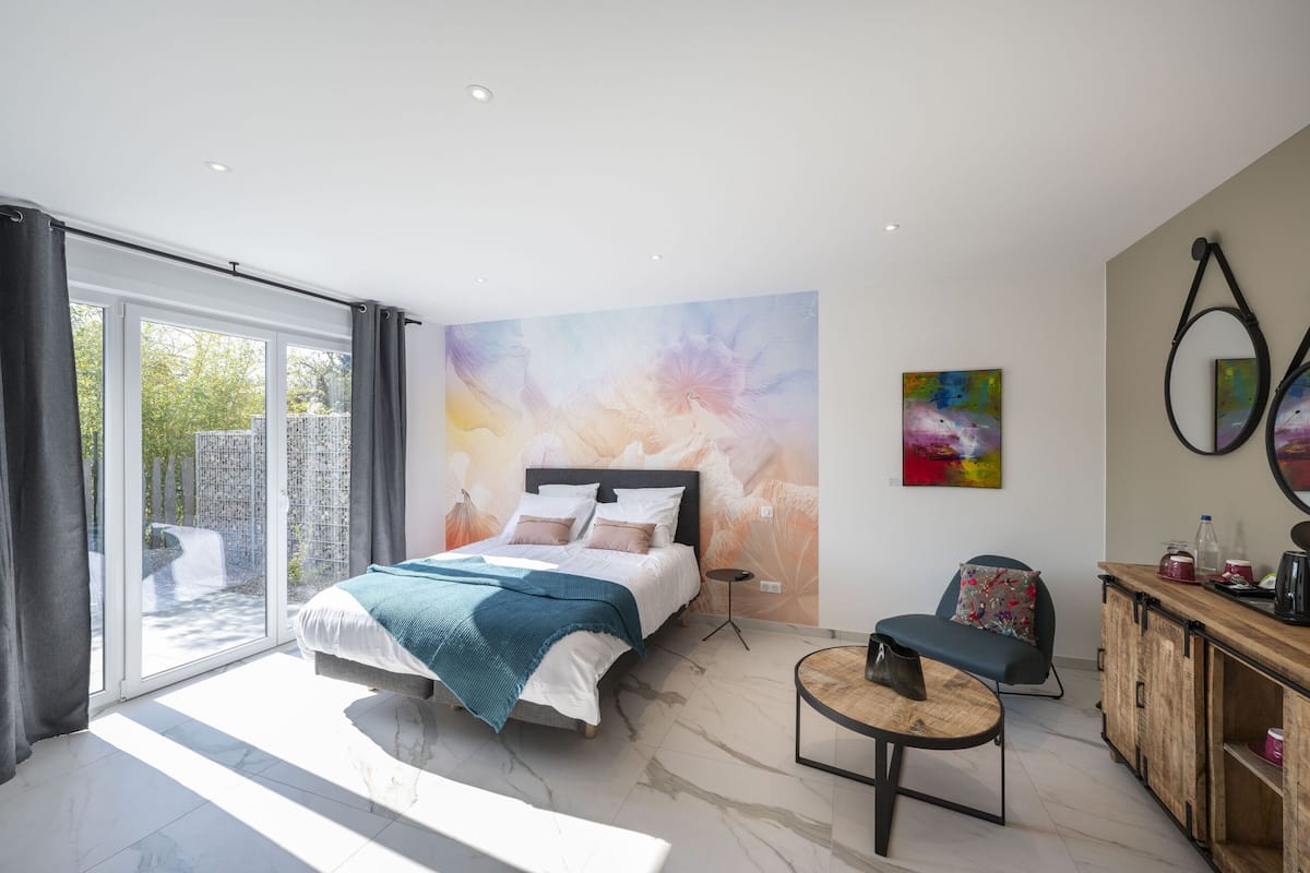 Ein modernes Schlafzimmer mit einem großen Bett, einem Schreibtisch und einem Sessel. Ein großes Glasfenster führt auf einen Balkon.
