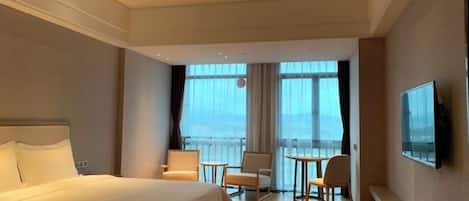 A hotel room with a large bed, a balcony with a view, a TV, and a seating area with chairs and a table.