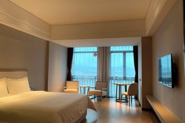 A hotel room with a large bed, a balcony with a view, a TV, and a seating area with chairs and a table.