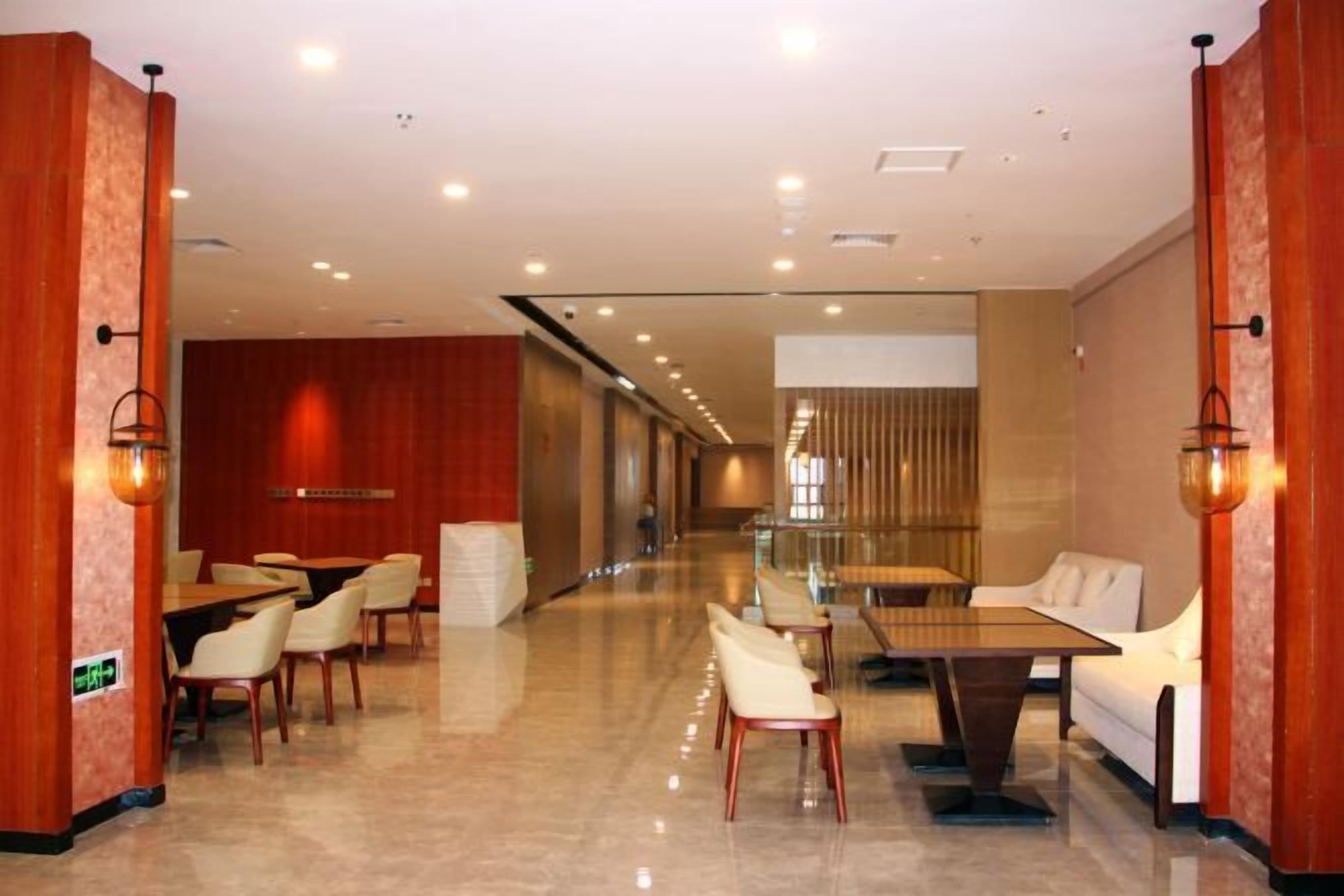 Un vestíbulo de hotel con una larga recepción, área de asientos con mesas y sillas, y un muro divisor de vidrio.