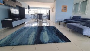 A modern living room with a large blue rug, a sofa, a TV, and a dining area in the background. - Appartement Duplex Bord de mer à Proximité de Tout ! (Saint-François)