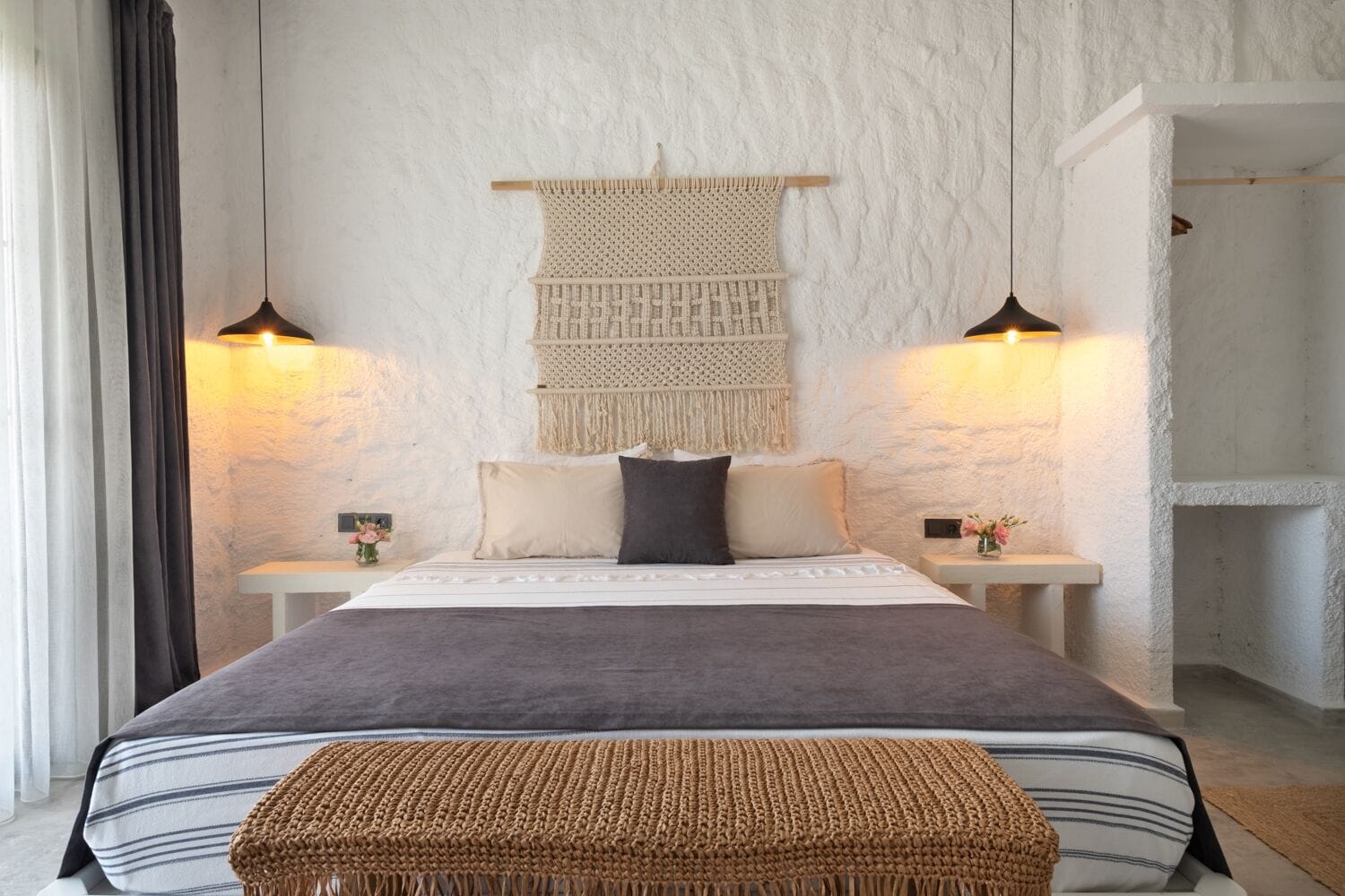 A bedroom with a bed, two bedside tables, a wall-mounted macrame piece, and two pendant lights.