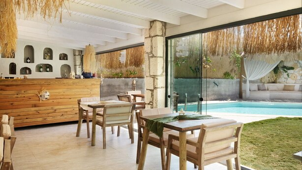A modern outdoor dining area with wooden tables and chairs, a stone wall with shelves, and a view of a swimming pool.