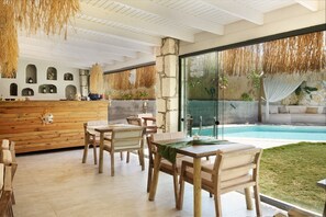 A modern outdoor dining area with wooden tables and chairs, a stone wall with shelves, and a view of a swimming pool.