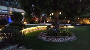 Un jardín bien iluminado con un sendero de piedra, un pequeño árbol y un edificio con techo de tejas al fondo.