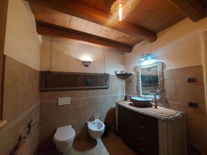 Un baño con techo de madera, inodoro blanco, bidet, tocador con espejo y repisa sobre el lavabo.